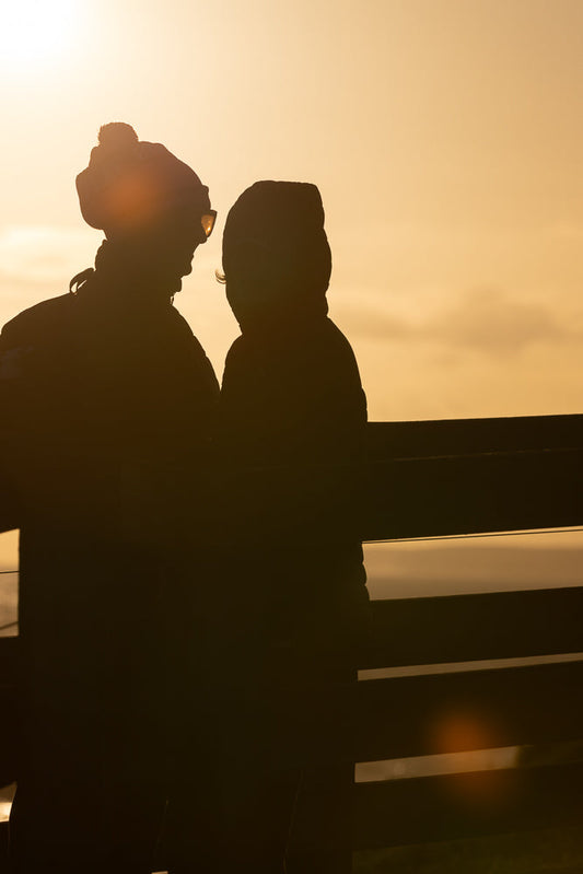 Silhouettes of two people, possibly a couple, sitting close together on a bench against a bright, hazy sunset. The person on the left wears sunglasses and a beanie, while the person on the right is mostly obscured by shadow.