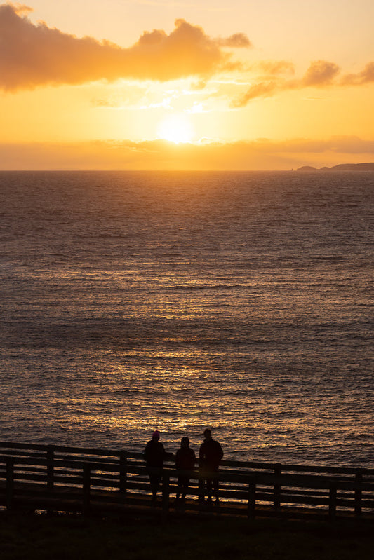 Three people stand on a wooden railing overlooking the ocean at sunset. The sun is setting on the horizon, casting a warm orange glow across the sky and reflecting on the water.