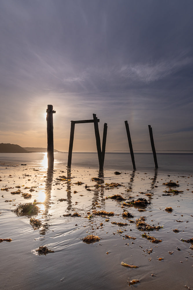 The sun sets behind the remnants of a wooden structure on a beach, casting long reflections on the wet sand. The sky is filled with wispy clouds, and the sea is calm.