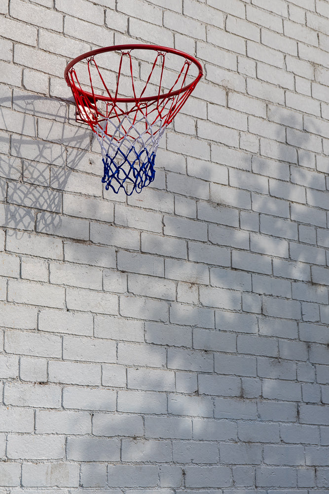 A basketball hoop with a red rim and a red, white, and blue net is mounted on a white brick wall. Shadows of leaves are cast on the wall.