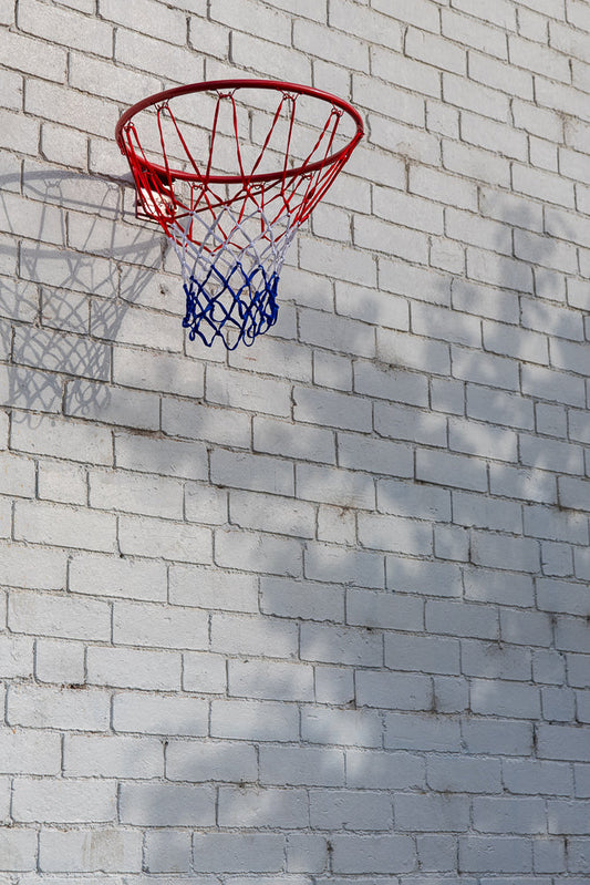 A basketball hoop with a red rim and a red, white, and blue net is mounted on a white brick wall. Shadows of leaves are cast on the wall.