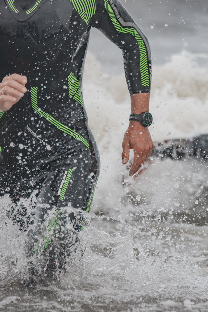 A close-up shot of a triathlete in a black and green wetsuit running through water, with splashes and droplets visible.