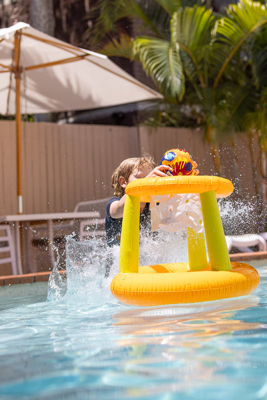 A young boy with blond hair is playing with a colorful inflatable basketball hoop in a swimming pool. He is splashing water as he reaches for the ball.