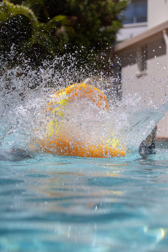 A bright orange and yellow water balloon explodes in a splash of water in a swimming pool. The image is taken from a low angle, showing the water balloon breaking apart and water spraying everywhere.