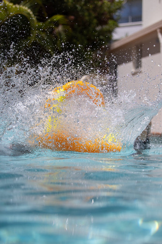 A bright orange and yellow water balloon explodes in a splash of water in a swimming pool. The image is taken from a low angle, showing the water balloon breaking apart and water spraying everywhere.