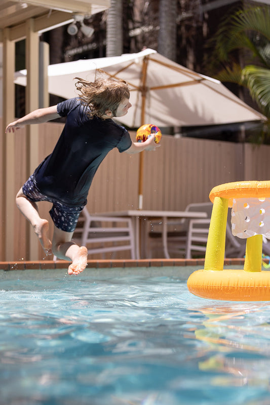 A young boy with wet hair jumps into a swimming pool, holding a colorful ball, aiming for an inflatable basketball hoop.