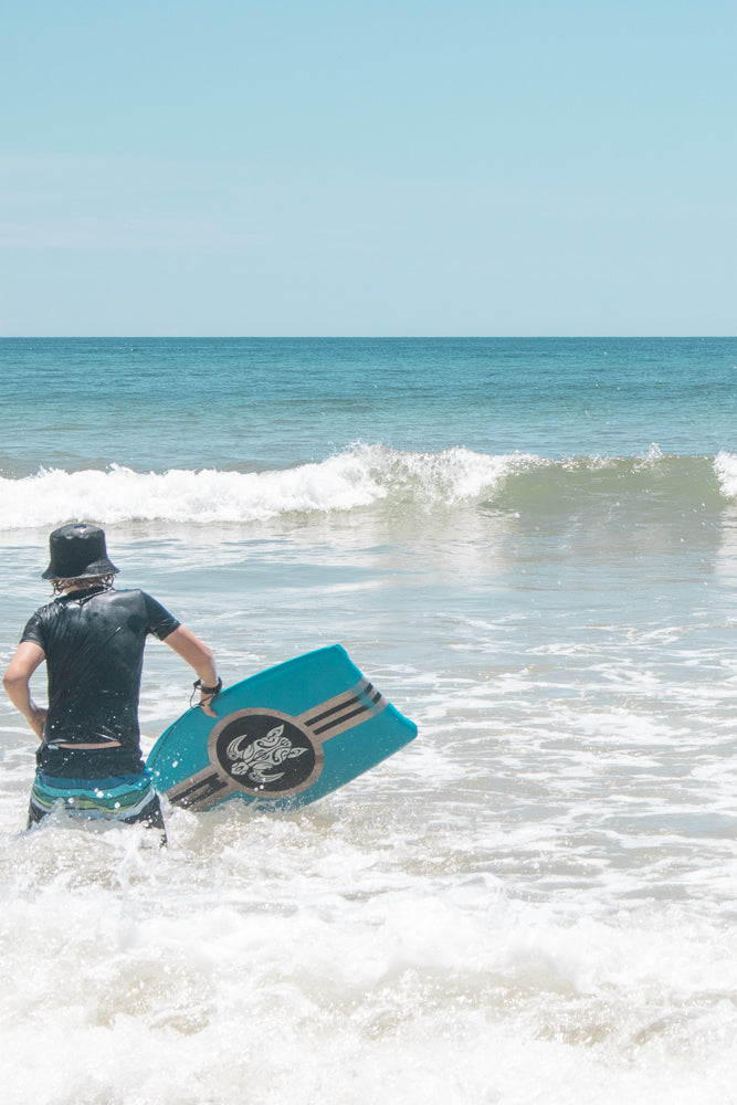 A person wearing a black shirt and striped shorts walks into the ocean holding a blue boogie board. The water is choppy with white foam.