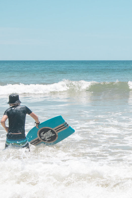 A person wearing a black shirt and striped shorts walks into the ocean holding a blue boogie board. The water is choppy with white foam.