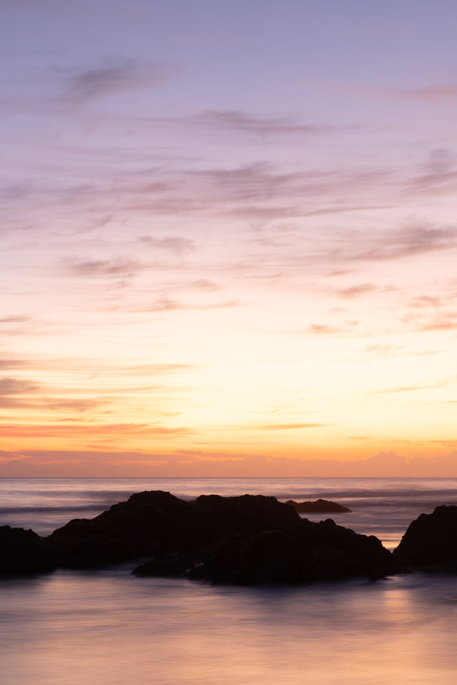 A serene seascape at dusk, with dark, silhouetted rocks in the foreground against a soft, pastel-colored sky transitioning from pale yellow to lavender. The ocean water is smooth and reflective, creating a dreamlike atmosphere.