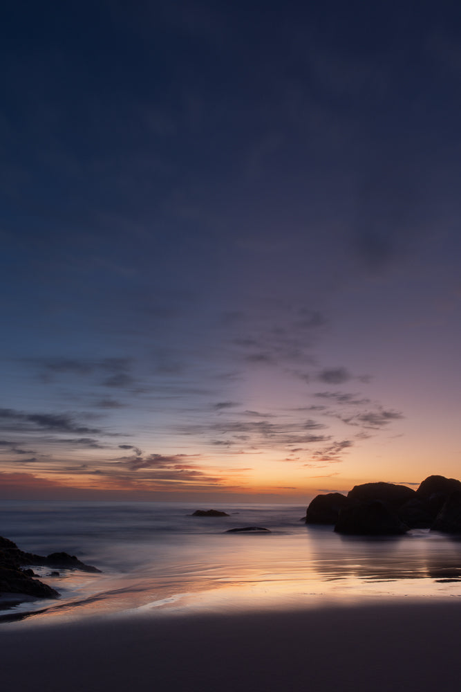 A serene beach scene at dusk. The sky transitions from deep blue to soft purple and orange hues, with wispy clouds. Smooth, reflective water laps at the shore, with dark, silhouetted rocks scattered in the distance and foreground. The wet sand mirrors the colors of the sky.