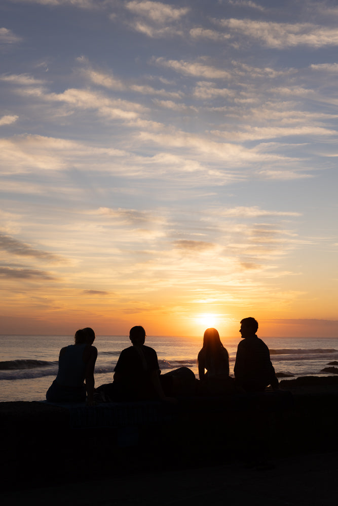 Four friends silhouetted against a vibrant sunset over the ocean, sitting on a rocky shore and watching the sun dip below the horizon.