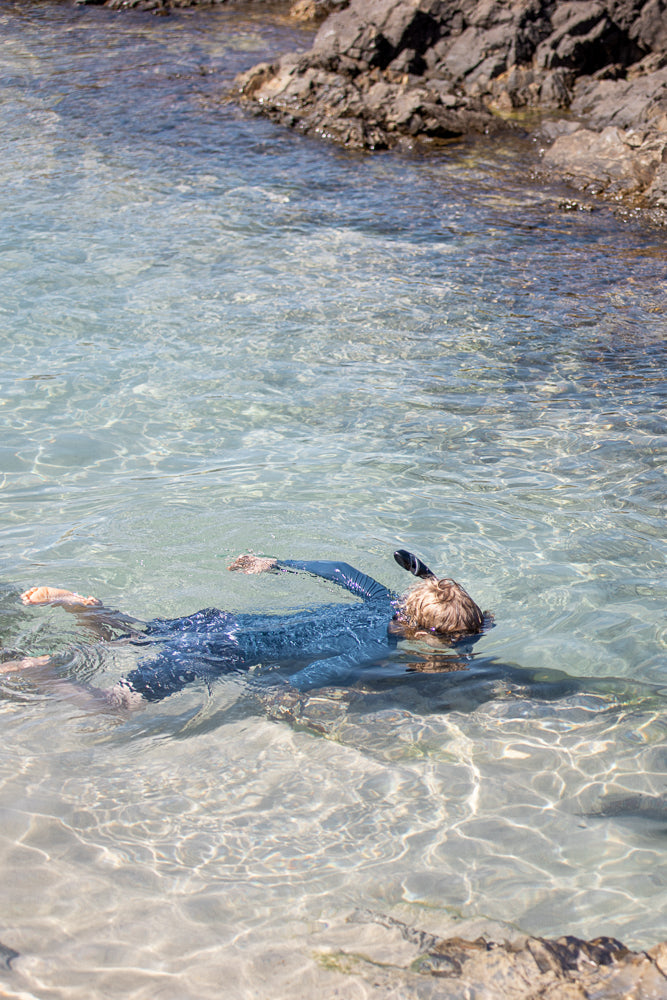 A young person wearing a blue rash guard and snorkel mask floats on their back in clear, shallow water near rocky shore.