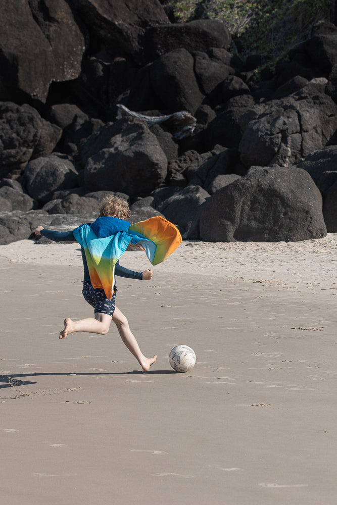 A young boy with blond hair runs barefoot on a sandy beach towards a soccer ball. He wears a dark blue long-sleeved shirt and patterned shorts, with a colorful towel draped over his shoulders.