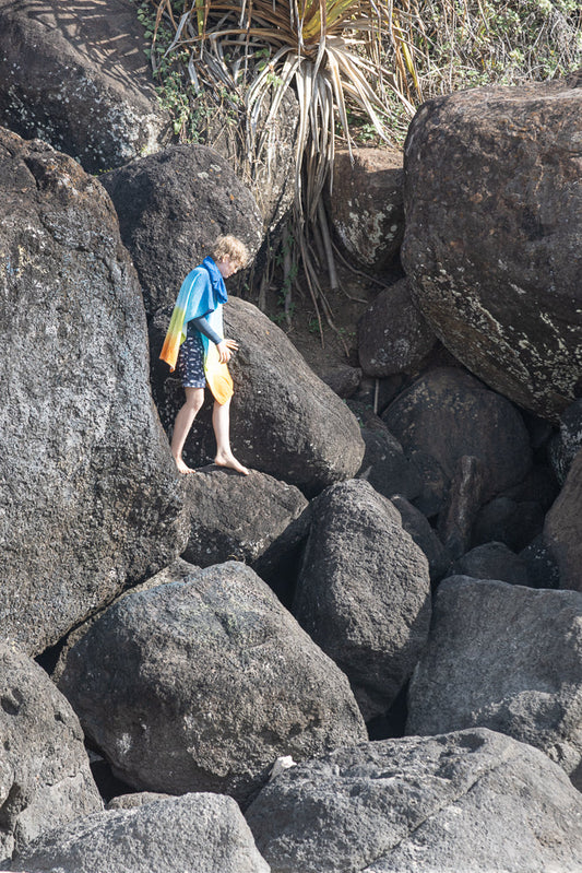A young boy with curly blonde hair walks barefoot across large, dark rocks. He wears a blue and yellow tie-dye towel draped over his shoulders and patterned swim trunks. He holds a yellow object in his right hand.