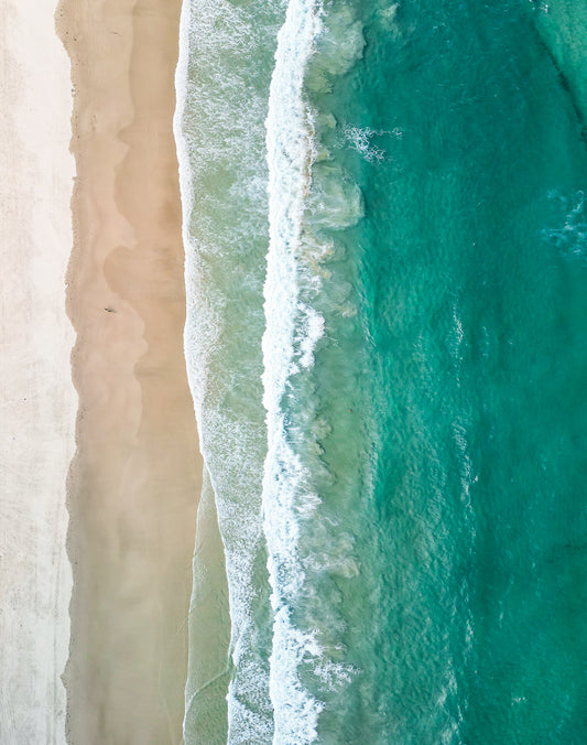 Aerial view of a sandy beach with gentle waves rolling in. The water is a clear turquoise color, transitioning to lighter shades near the shore. The sand is a light beige, with some footprints visible.