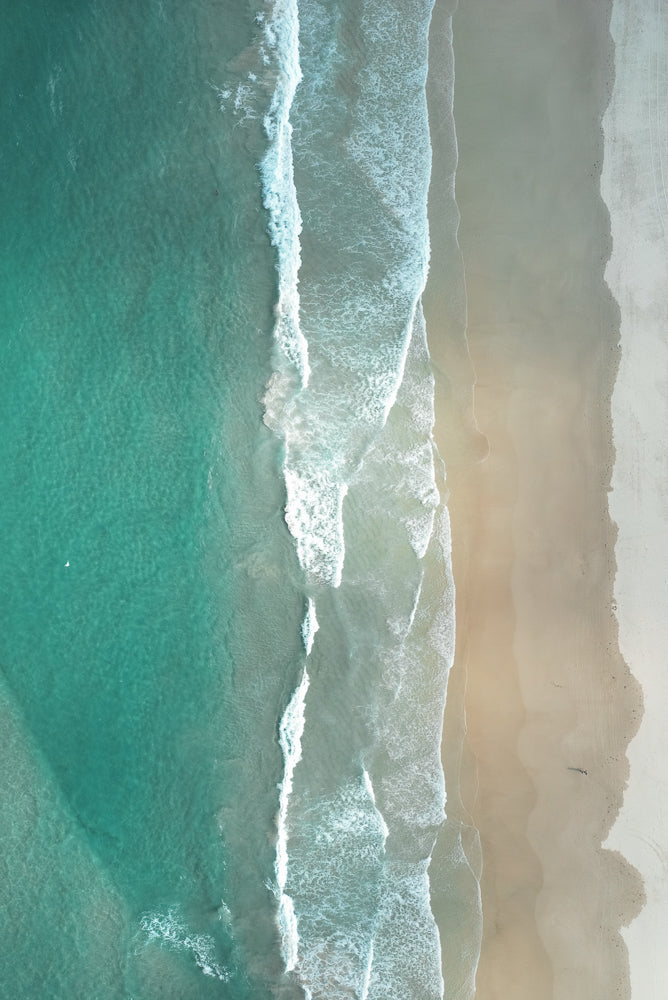 An aerial view of a beach shows the turquoise ocean meeting the sandy shore with gentle waves rolling in. A lone figure walks along the wet sand near the water's edge.