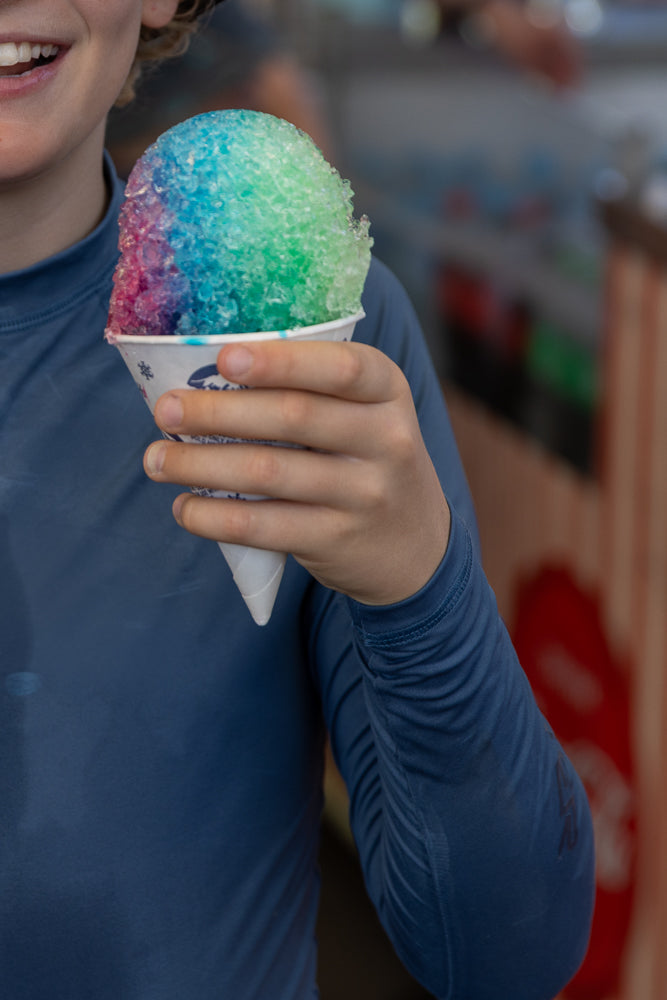 A person in a blue shirt holds a cup of rainbow-colored shaved ice, with their mouth visible and smiling.