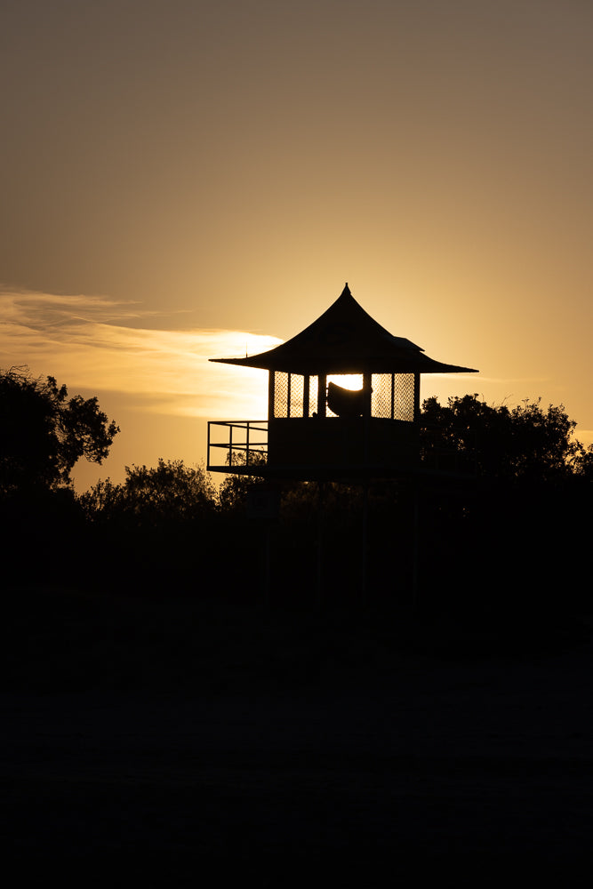 Silhouette of a lifeguard tower against a golden sunset sky. The tower has a distinctive pagoda-style roof and a railing around its platform. Trees are silhouetted in the foreground and background.