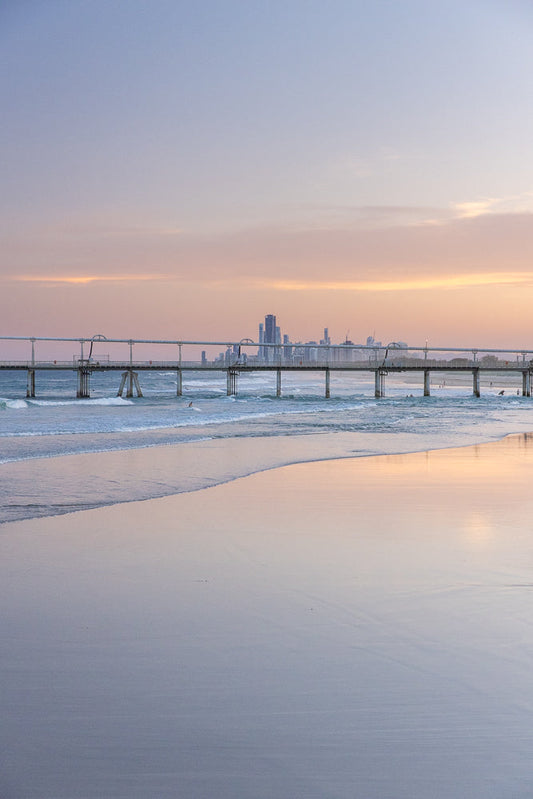A wide shot of a beach at sunset with a pier extending into the ocean. The sky is filled with soft pink and purple hues, reflecting on the wet sand. Gentle waves roll onto the shore, and a few surfers can be seen in the water.