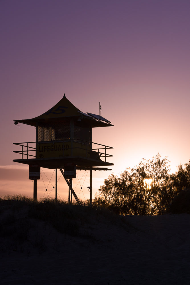 Silhouette of a lifeguard tower on a beach at sunset. The sky is a gradient of purple and pink, with the sun peeking through the trees behind the tower.