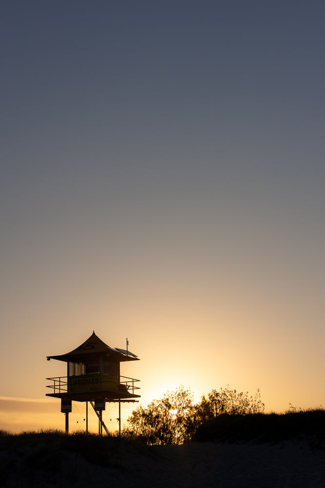 A silhouette of a lifeguard tower stands against a golden sunset sky. The sun is setting behind a cluster of trees, casting a warm glow across the scene.