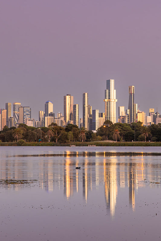 A rowing crew practices on a calm lake in front of the Melbourne skyline at sunrise.