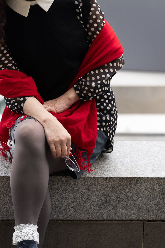 A woman sits on a granite ledge, wearing a black top with white polka dots, a red scarf, and grey tights with white lace trim around her ankles. She holds a pair of glasses in her hand.