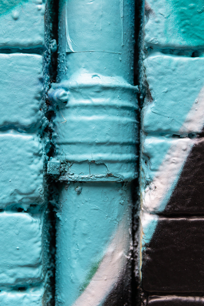 A close-up, abstract shot of a pipe covered in textured turquoise paint, set against a brick wall also painted turquoise. The pipe has horizontal ridges and shows signs of cracking and wear. To the right, the wall is painted with bold diagonal stripes of black and white.