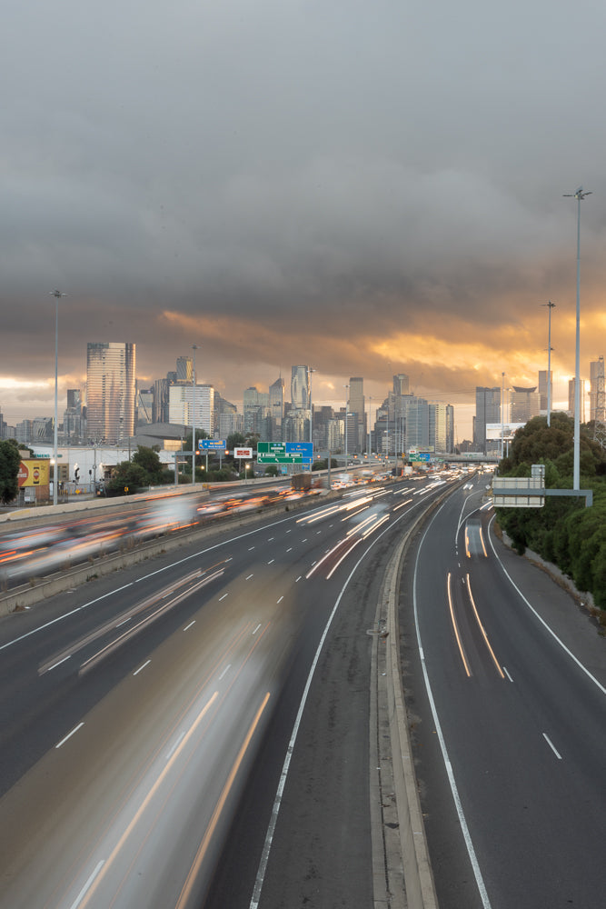 A long exposure shot of a highway with light trails from cars, leading towards a city skyline under a dramatic, cloudy sky with golden sunset light breaking through.