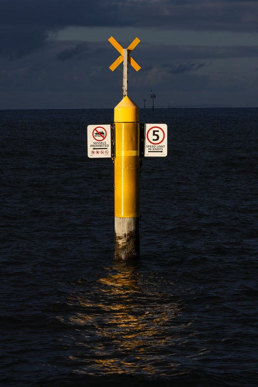 A yellow navigational buoy stands in dark water under a cloudy sky. The buoy has an X-shaped marker at the top and two signs attached to its side. One sign indicates "Vessels Prohibited" with an arrow pointing left, and the other indicates a "5 Speed Limit in Knots" with an arrow pointing right.