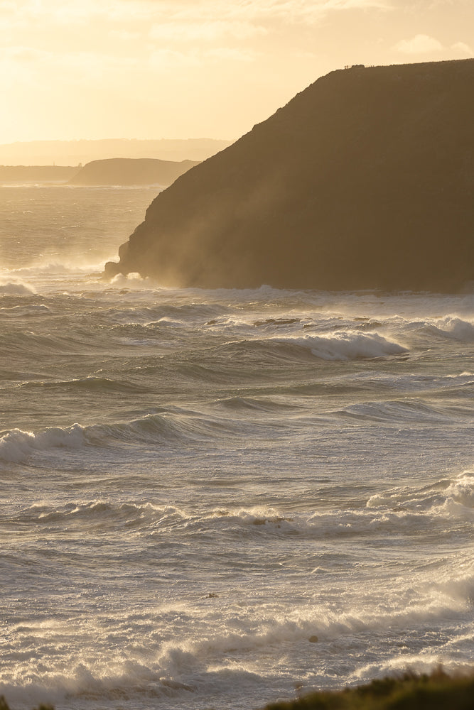 A dramatic coastal scene at sunset with rough, choppy waves crashing against a dark, silhouetted cliff face. The golden light of the setting sun illuminates the spray and foam, creating a powerful and atmospheric view.