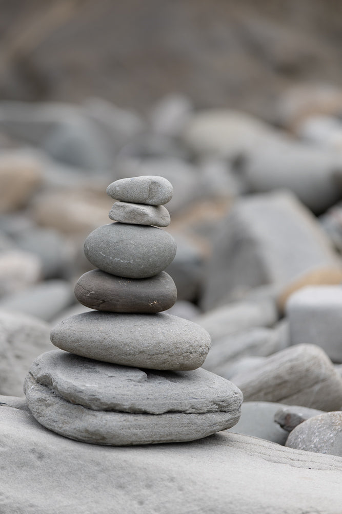 A stack of smooth, grey stones balanced on a rocky shore. The stones are of varying sizes, with the largest at the bottom and the smallest at the top. The background is blurred, showing more rocks and a muted, natural environment.
