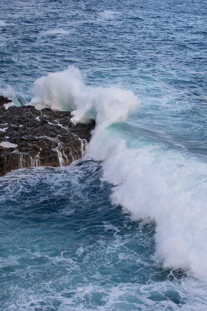 A powerful wave crashes against dark, jagged rocks on a coastline. The water is a deep blue, with white foam and spray erupting from the impact.
