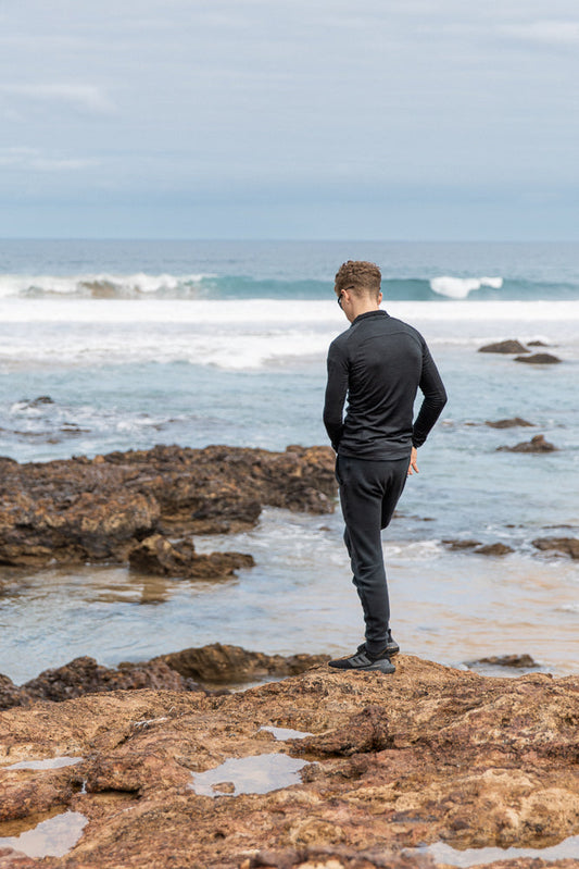 A young man with curly hair stands on a rocky shore, looking down at the ocean. Waves crash in the background under a cloudy sky.