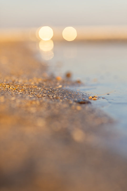 A close-up, low-angle shot of a sandy beach with small pebbles and shells at the edge of the water. The background is blurred with soft, golden bokeh lights.