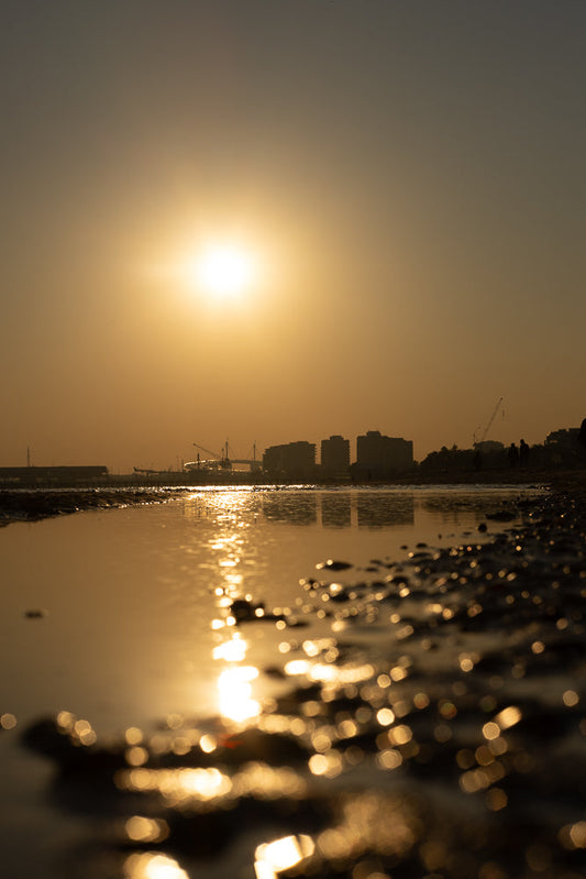 The sun sets over a calm body of water, reflecting the golden light and the silhouettes of buildings and cranes in the distance. The foreground is filled with bokeh lights from the water's surface.