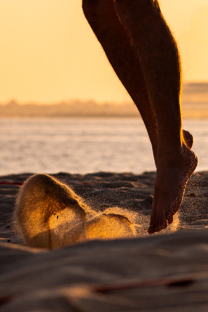 A person's legs and feet are shown in silhouette against a golden sunset sky as they run on a beach. Sand is kicked up around their feet, sparkling in the light.