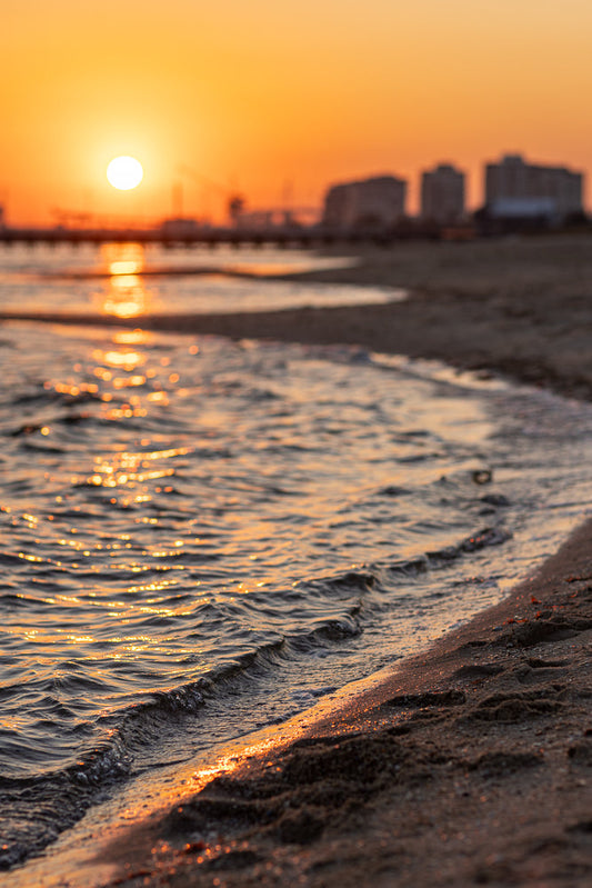 A serene beach scene at sunset. The sun, a bright orb, hangs low in the orange sky, casting a shimmering golden path across the gentle waves. The water laps at the sandy shore, reflecting the warm light. In the blurred background, a distant cityscape is visible.
