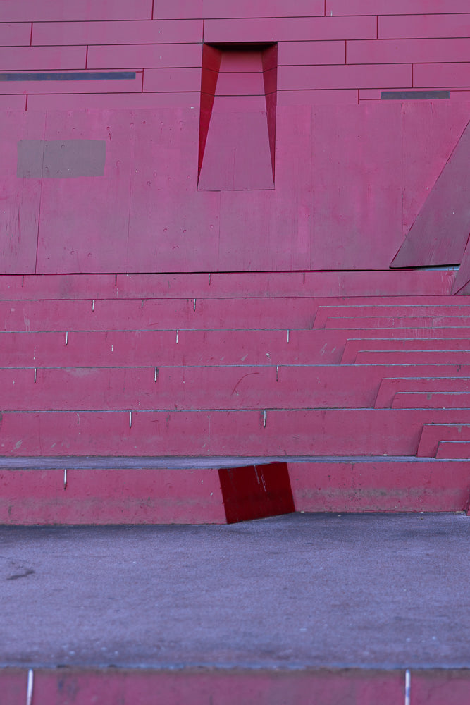A close-up, abstract view of a large, modern, red structure with horizontal lines and geometric cutouts. The lower portion shows tiered steps leading to a grey ground surface.