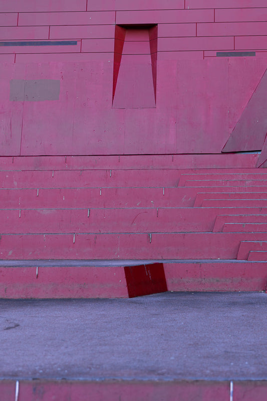 A close-up, abstract view of a large, modern, red structure with horizontal lines and geometric cutouts. The lower portion shows tiered steps leading to a grey ground surface.