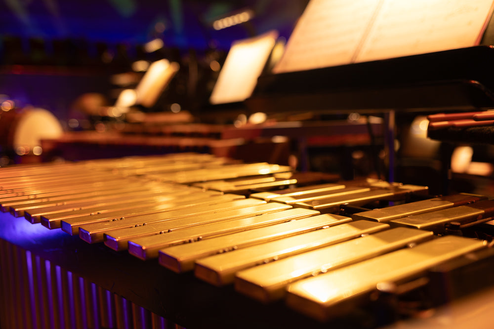 A close-up, low-angle shot of a xylophone with golden bars, illuminated by purple and blue stage lights. The instrument is in focus in the foreground, with blurred musical instruments and sheet music stands in the background.
