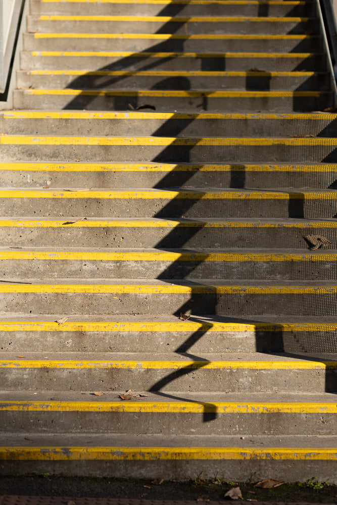 A close-up view of concrete stairs with bright yellow safety strips on each step. A sharp, angular shadow from a railing or fence falls across the stairs, creating a geometric pattern of light and dark.