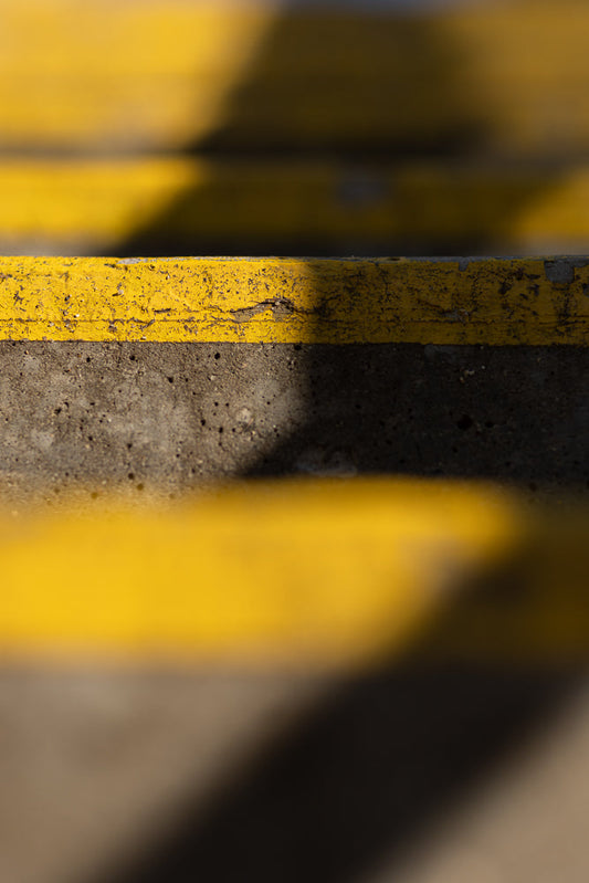 A close-up, abstract shot of concrete steps with bright yellow safety stripes. The stripes are worn and chipped, showing the texture of the concrete beneath. Shadows create a dramatic effect across the steps.
