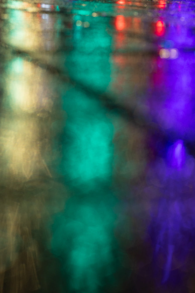 Abstract bokeh of colorful lights reflecting on a wet surface at night. The image features streaks of green, purple, and yellow light, creating a dreamy and atmospheric effect.
