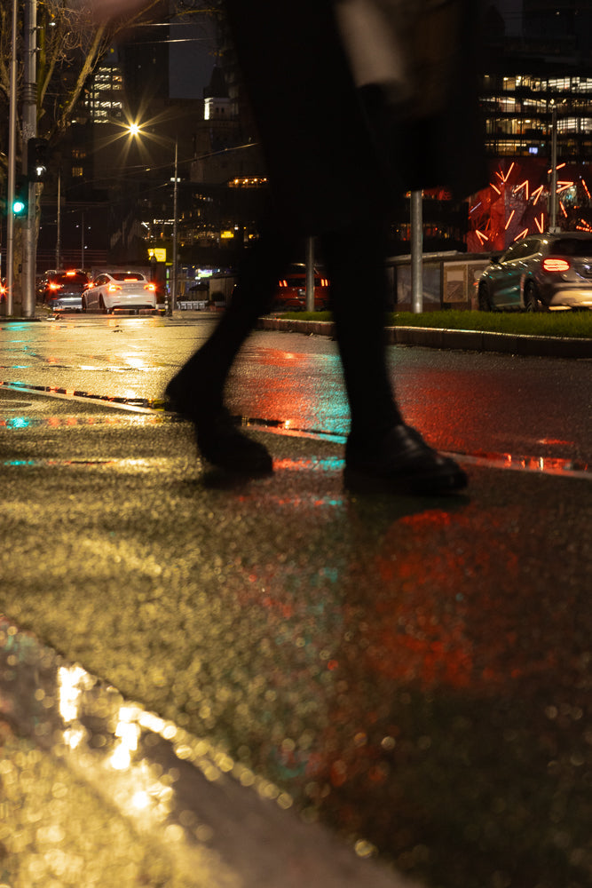 A person walks across a wet city street at night, with reflections of traffic lights and buildings in the puddles.