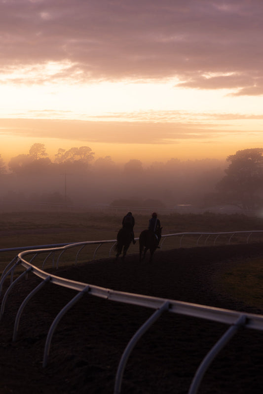 Two jockeys on horseback gallop along a misty racetrack at sunrise. The sky is a soft gradient of purple and orange, with clouds illuminated by the rising sun. Trees are silhouetted in the distance through the fog.
