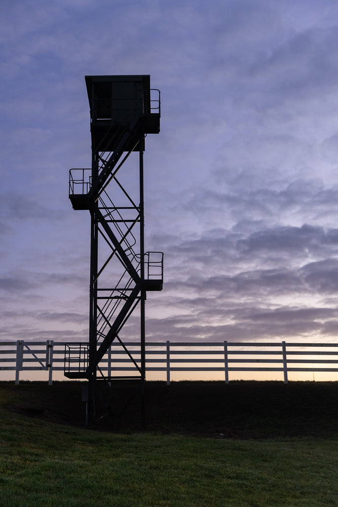 Silhouette of a tall observation tower with a staircase against a cloudy twilight sky. A white fence runs horizontally in the mid-ground.