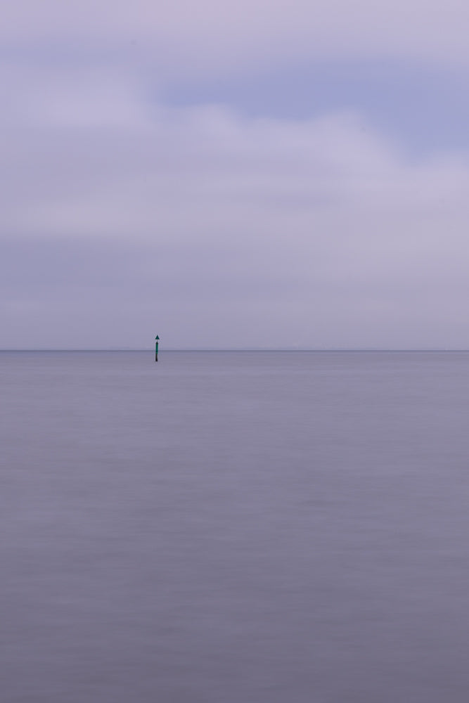A solitary green and black buoy stands in the vast, calm, and misty sea under a pale purple sky. The water is smooth and blurred, creating a serene and minimalist atmosphere.