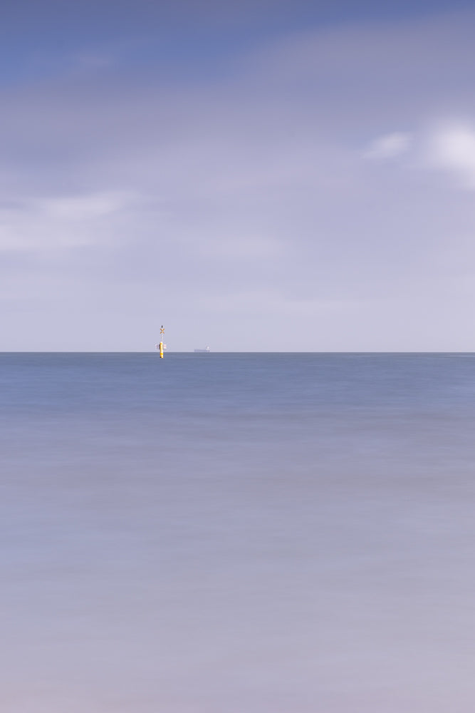 A yellow and white buoy with a cross on top stands in the calm blue ocean. In the distance, a large ship is visible on the horizon under a pale purple sky with wispy clouds.
