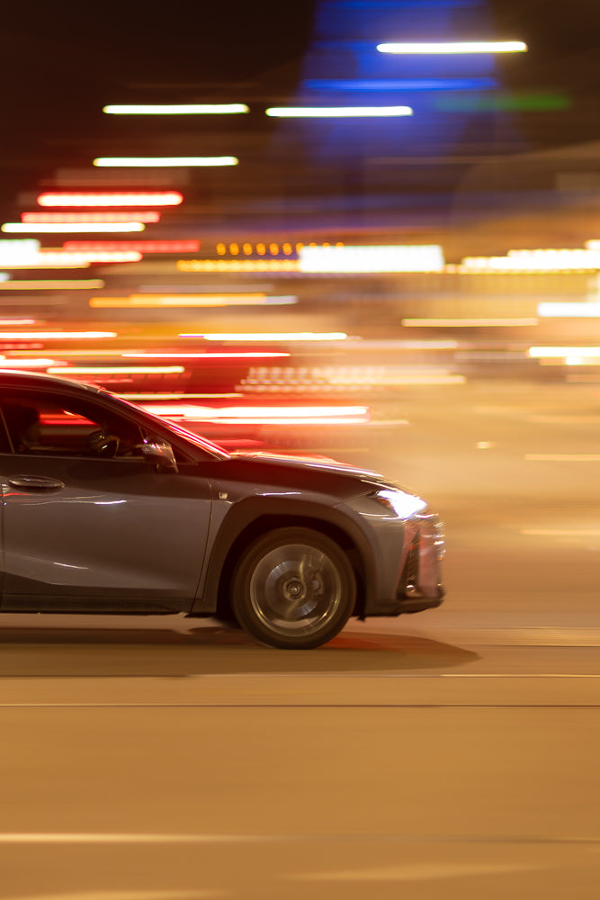 A grey car is driving at night, with streaks of red, white, and blue lights blurring past it, indicating motion.